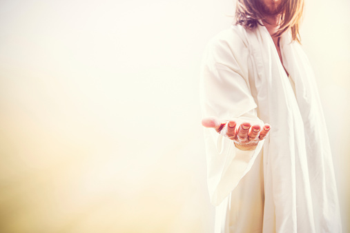 A representation of Jesus Christ following the resurrection in a heaven like setting, extending his hand with the invitation of new life.  His face is obscured, and bright golden light shines around him. A fitting image for Christ rising from the dead as celebrated on Easter Sunday. Horizontal image with copy space.